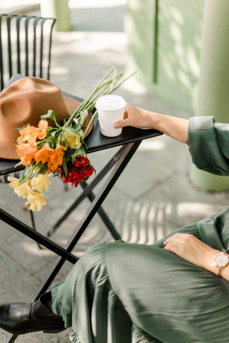 Eine Person, die mit Eleganz an einem Cafétisch sitzt, ihre Hand sanft auf einer Tasse Kaffee ruht, begleitet von einem lebendigen Blumenstrauß und einem stilvollen Hut daneben.