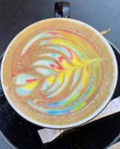 Eine farbenfrohe Latte Art mit einem lebendigen Regenbogendesign auf einer schaumigen Kaffeeleinwand, serviert in einer Keramiktasse, die dem Kaffeeerlebnis einen Hauch von Farben verleiht.