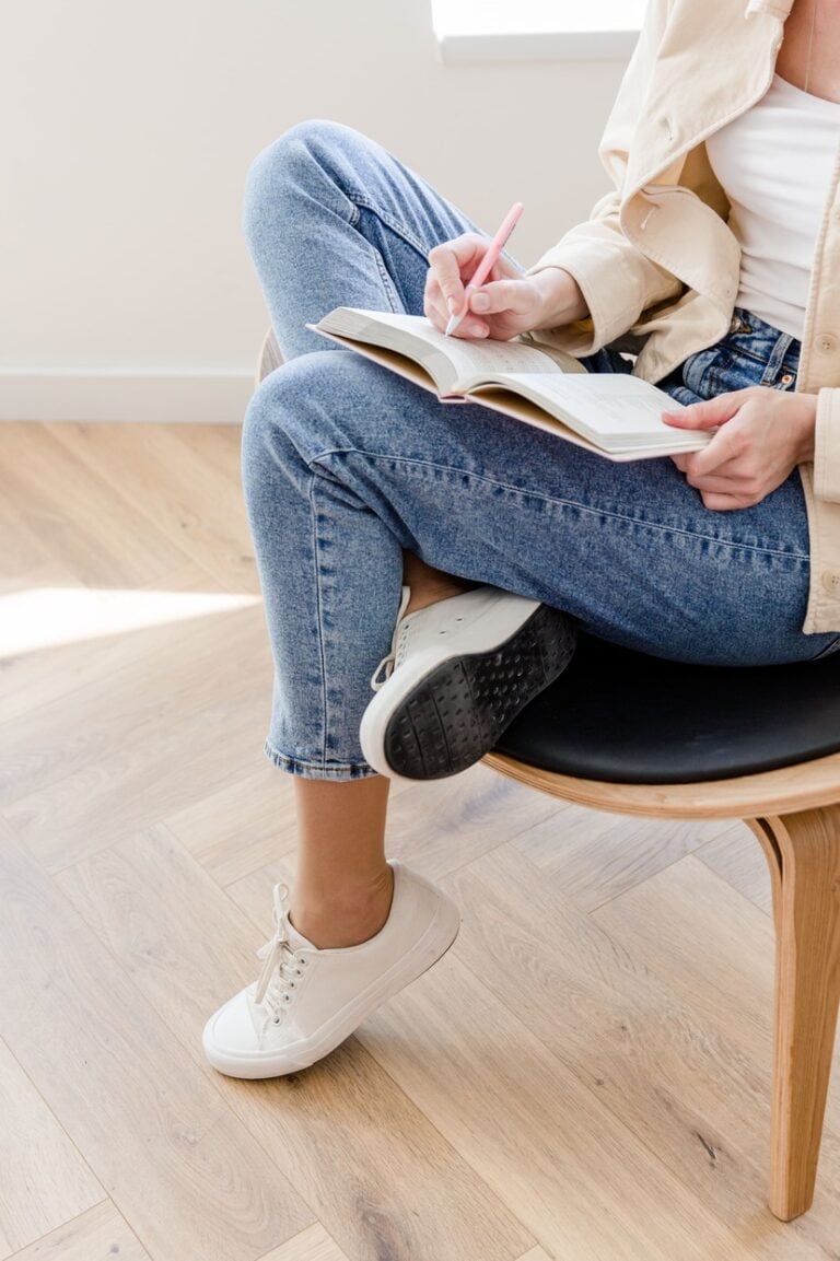 Eine Person in Jeans und weißen Turnschuhen sitzt auf einem Stuhl und macht sich Notizen in ein Notizbuch. Sie verkörpert ungezwungene Produktivität.