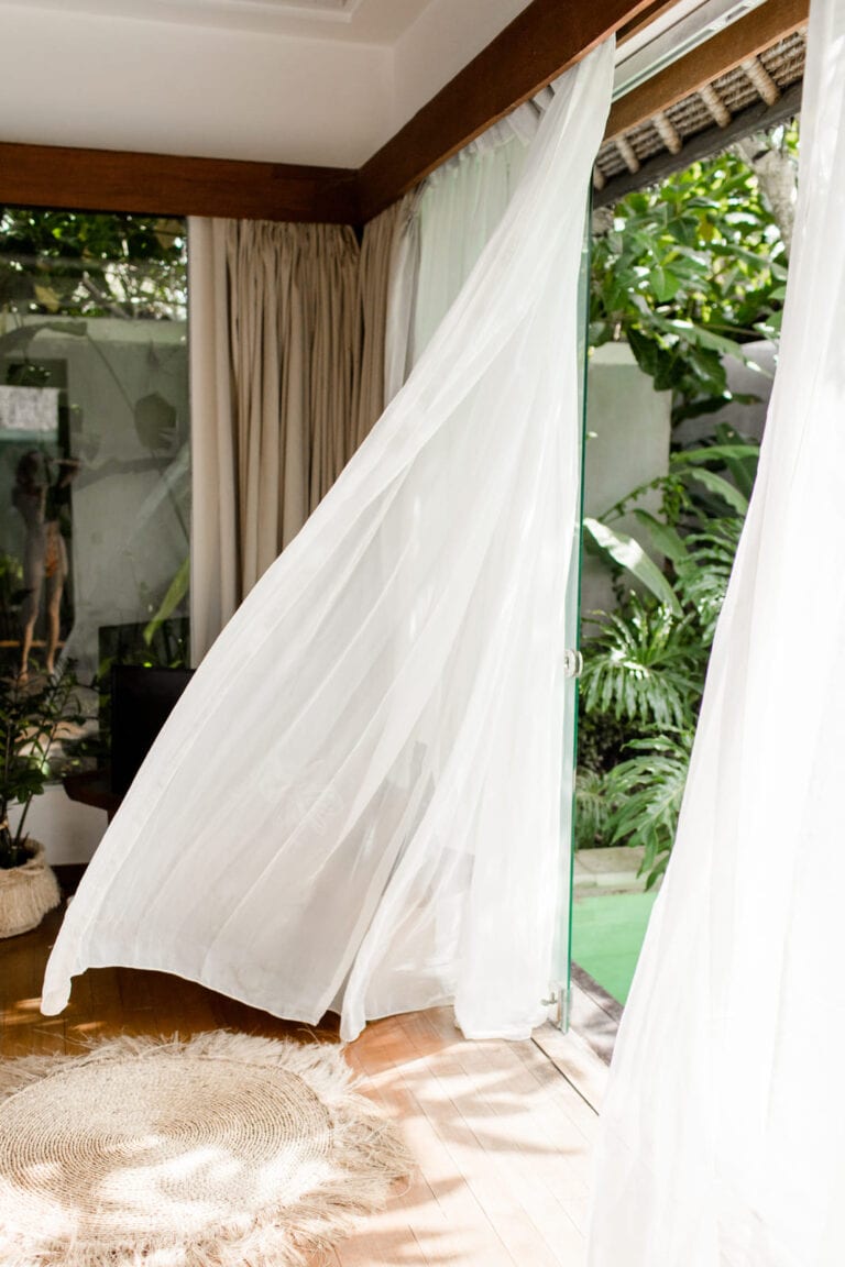 Ein luftiger und ruhiger Raum mit weißen, im Wind wehenden Vorhängen, der durch die offene Glastür in einen üppig grünen Garten führt zum Entspannen einlädt.