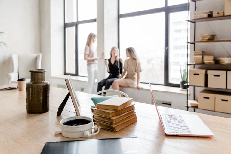 Ein gemütlicher Büroraum mit einem einladenden Schreibtisch im Vordergrund, auf dem ein Laptop, ein Stapel Bücher, eine Tasse Kaffee und ein Tablet und drei Frauen im Hintergrund.